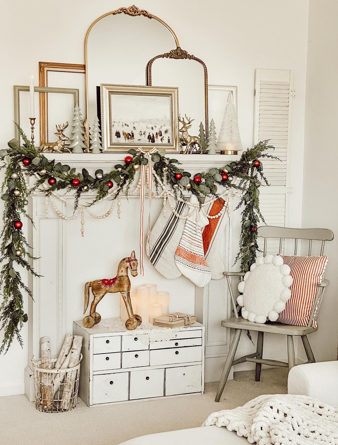 a white christmas mantle with antique art and mirrors, pine garland with silver and red ornaments, wooden beads, and small porcelain and gold holiday statues