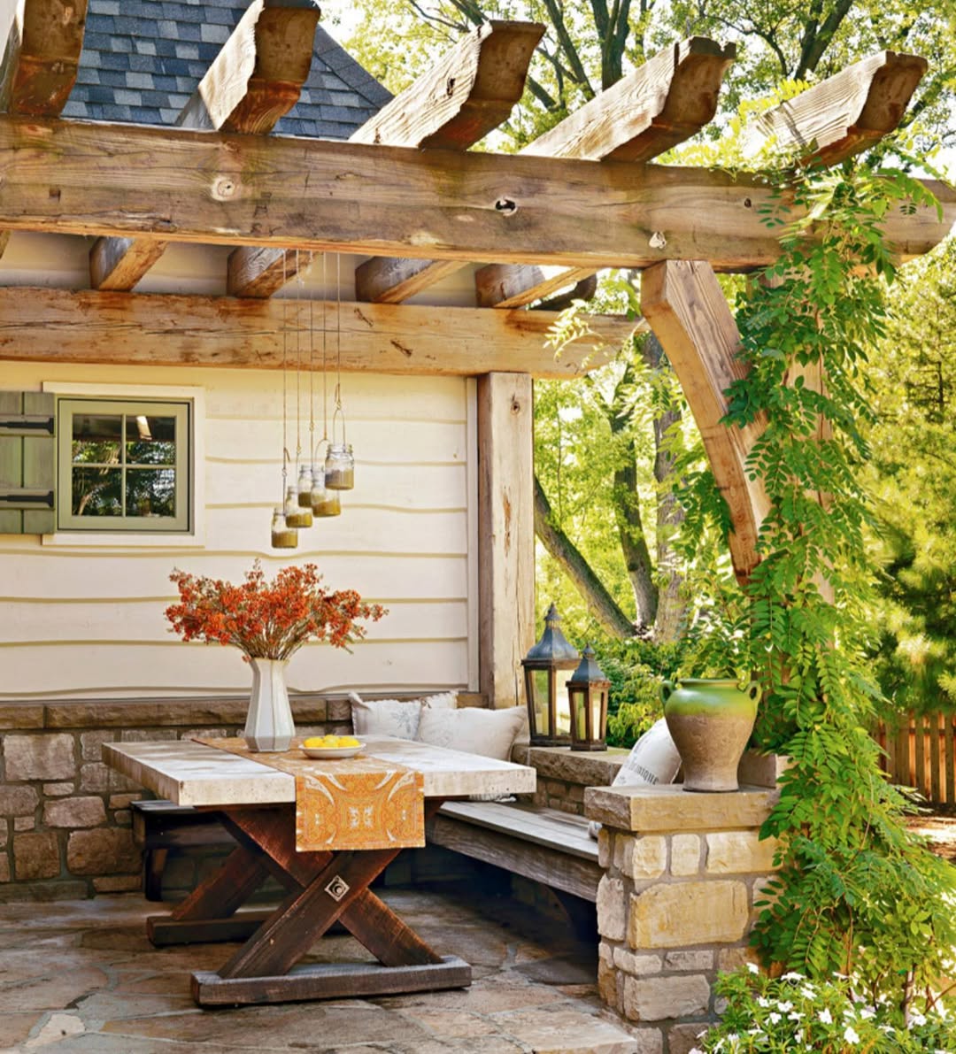 A rustic backyard patio featuring a wooden pergola, stone accents, picnic-style table, bench seating, hanging lanterns, and lush plants—perfect for outdoor destination inspiration and creative pergola ideas.